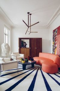 Salón con colores vivos en sus elementos. Sofá en naranja, alfombra con rayos en azul y fondo blanca, armario de madera, sillones en blanco.
